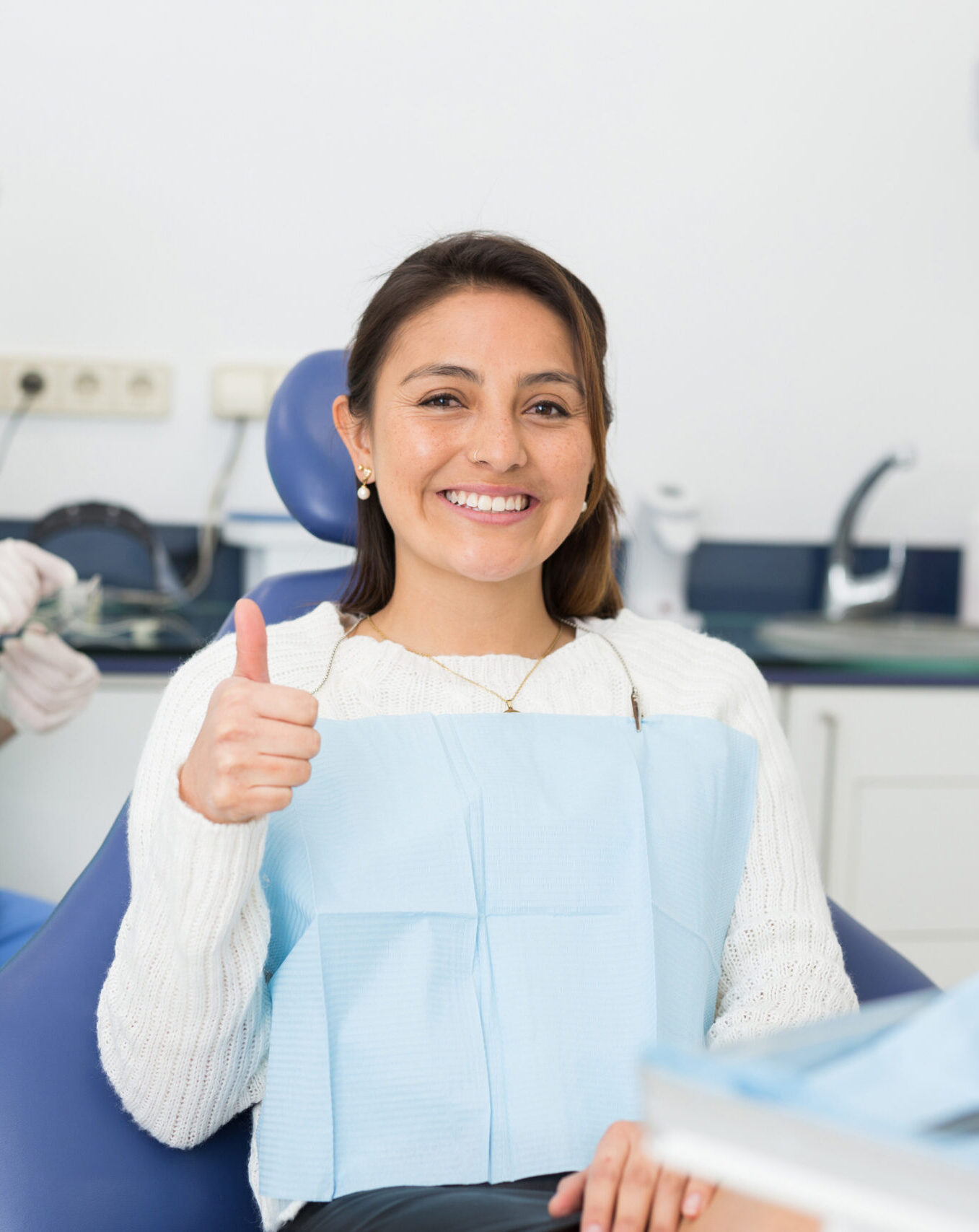 Happy cute latin woman sitting in dental chair after teeth cure giving thumb up